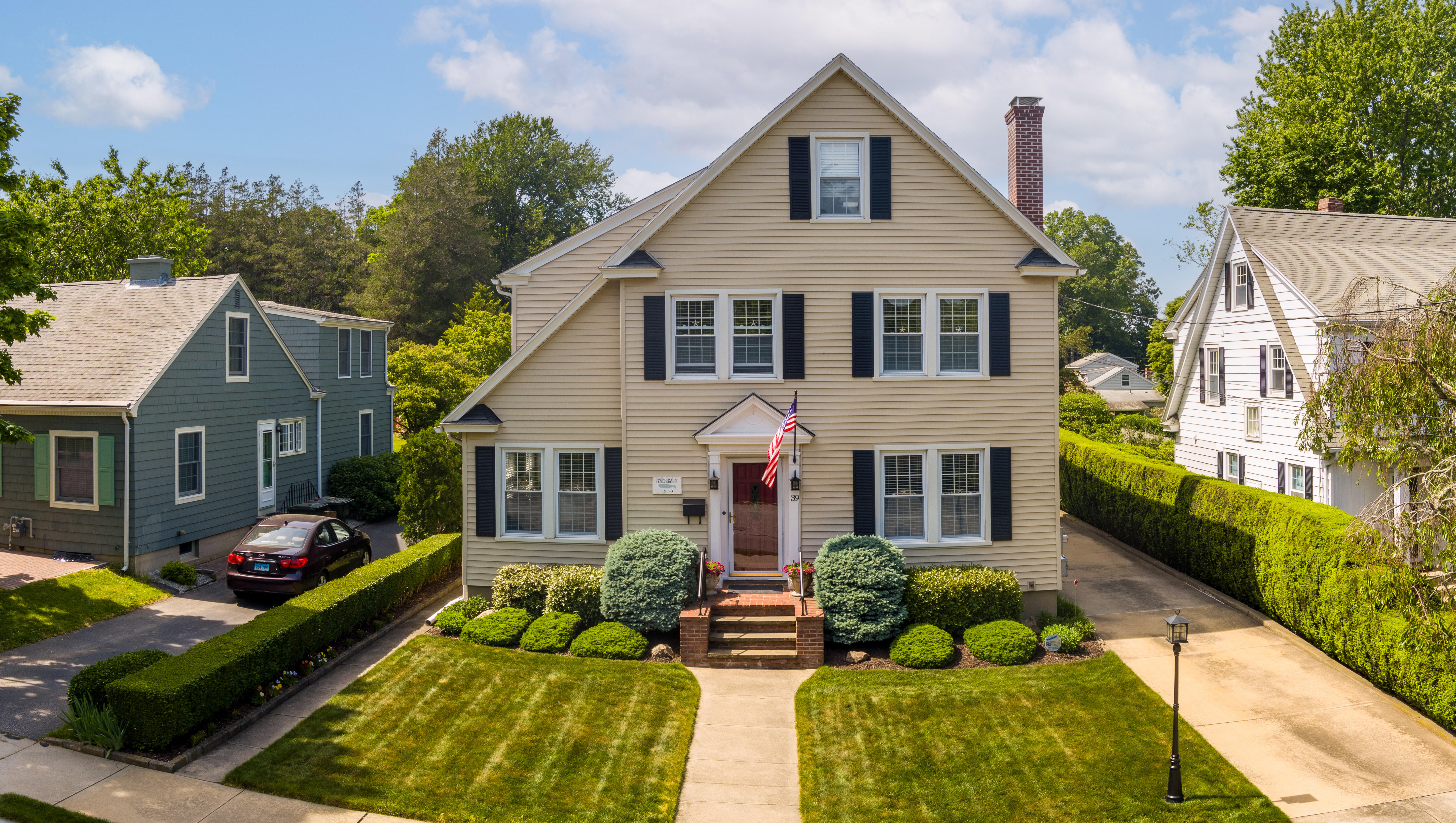 A two-story beige house with black shutters and a brick chimney is centered in the image. A well-kept lawn with shrubs surrounds the front. An American flag hangs by the entrance. A driveway with a black car is visible to the left.