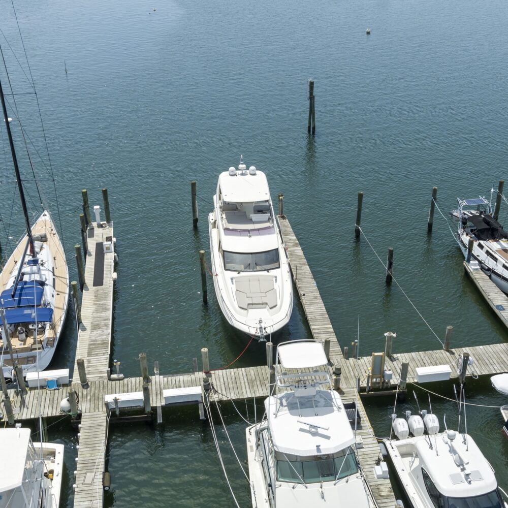 Aerial view of several boats and yachts docked at a wooden marina, with calm blue water and empty boat slips visible around the docks.