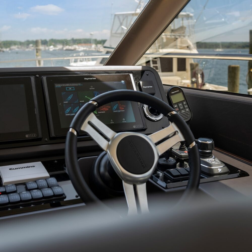 A close-up view of a yacht’s steering wheel and dashboard with navigation screens, controls, and a marine radio, docked at a marina with boats and water visible through the window.