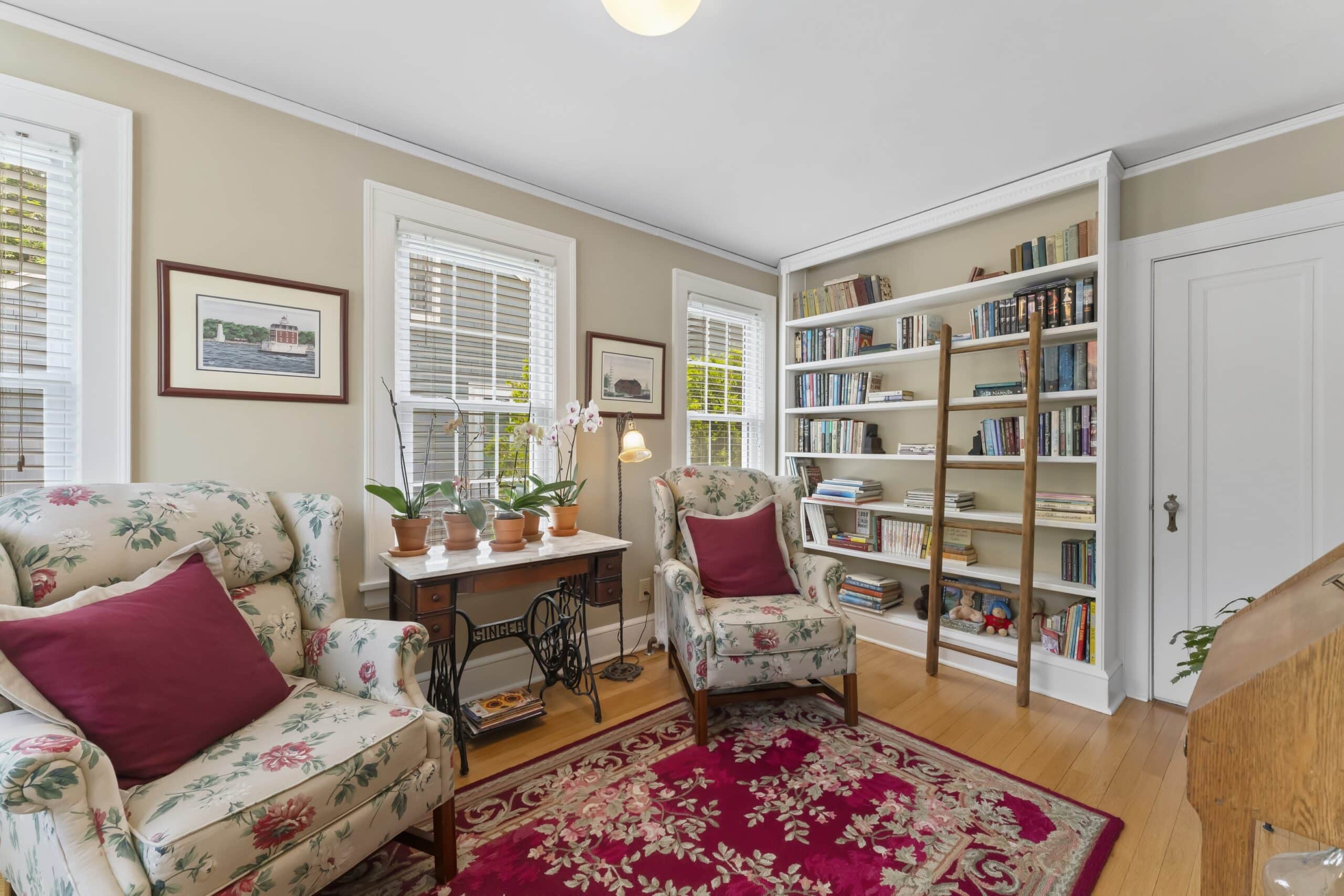 A cozy reading room with two floral armchairs, red accent pillows, and bookshelves comes to life in this image—perfect for showcasing through top-notch real estate media & photography services.
