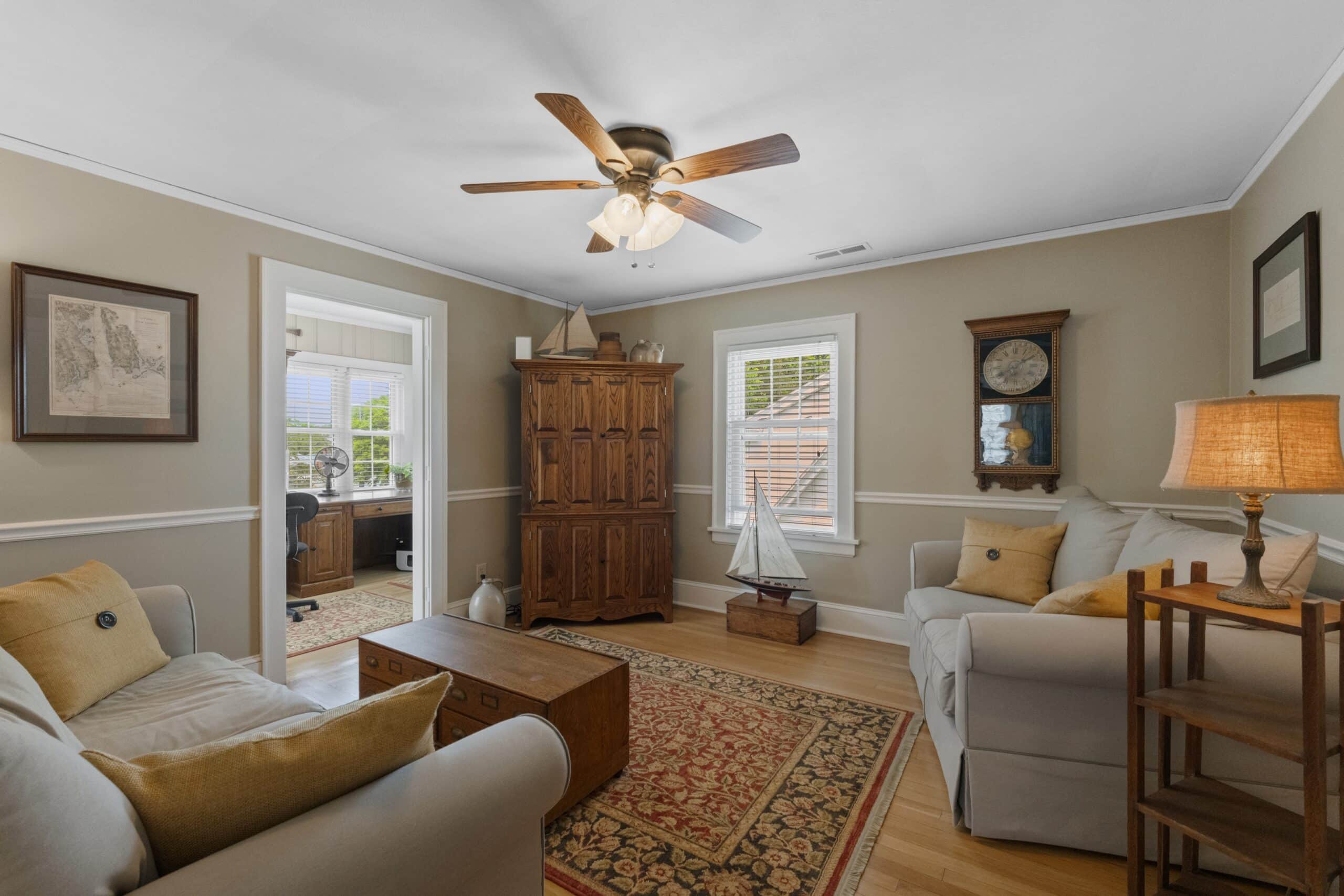 A cozy living room, ideal for Short Term Rental Photography in Waterford Connecticut, with beige walls, two sofas, a wooden chest coffee table, a patterned rug, large armoire, ceiling fan, window, and doorway to a sunlit office area.