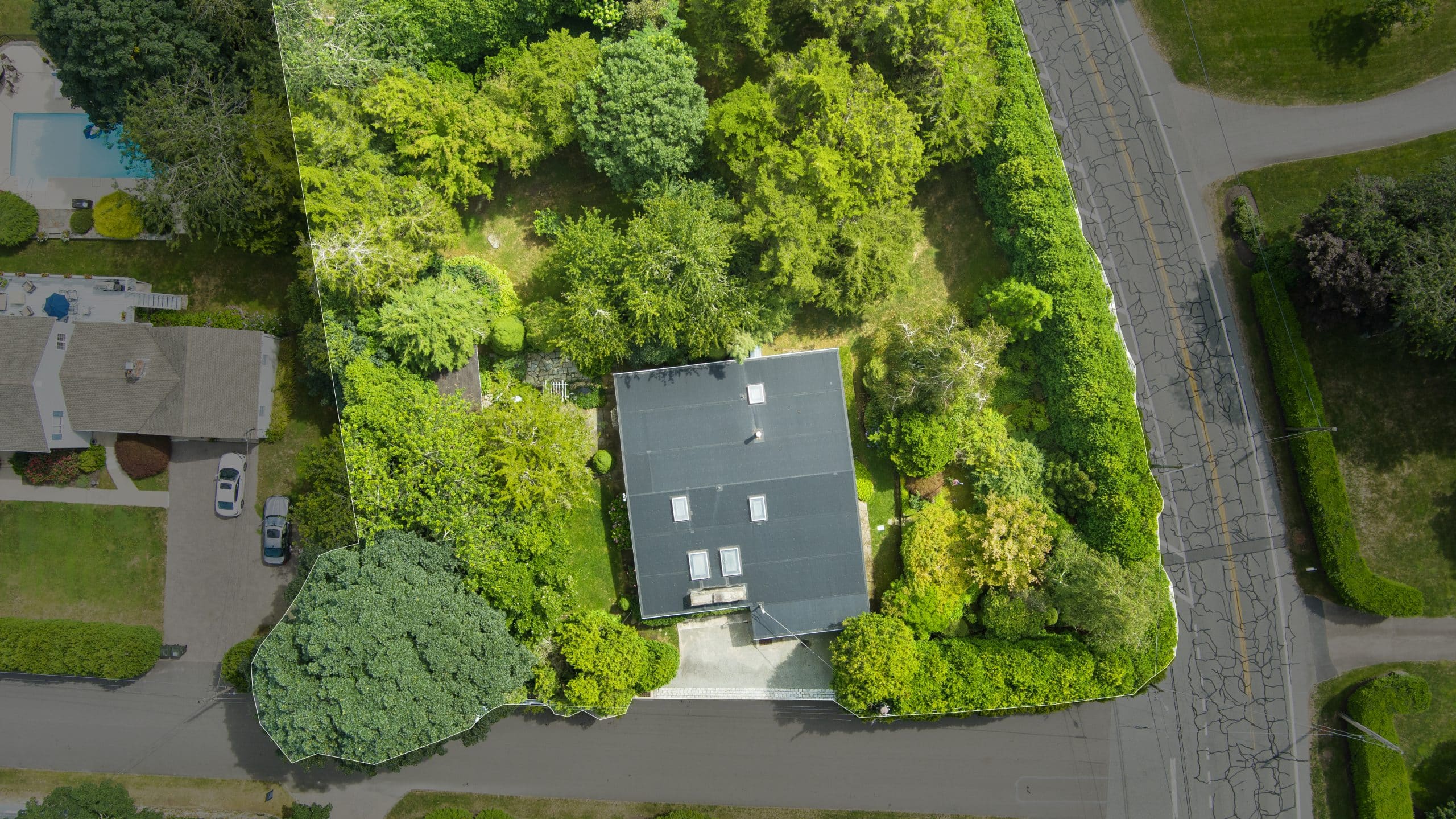 Aerial view of a home with a dark roof surrounded by lush green trees and gardens. The property is bordered by roads on three sides, and other houses and a swimming pool are visible nearby.