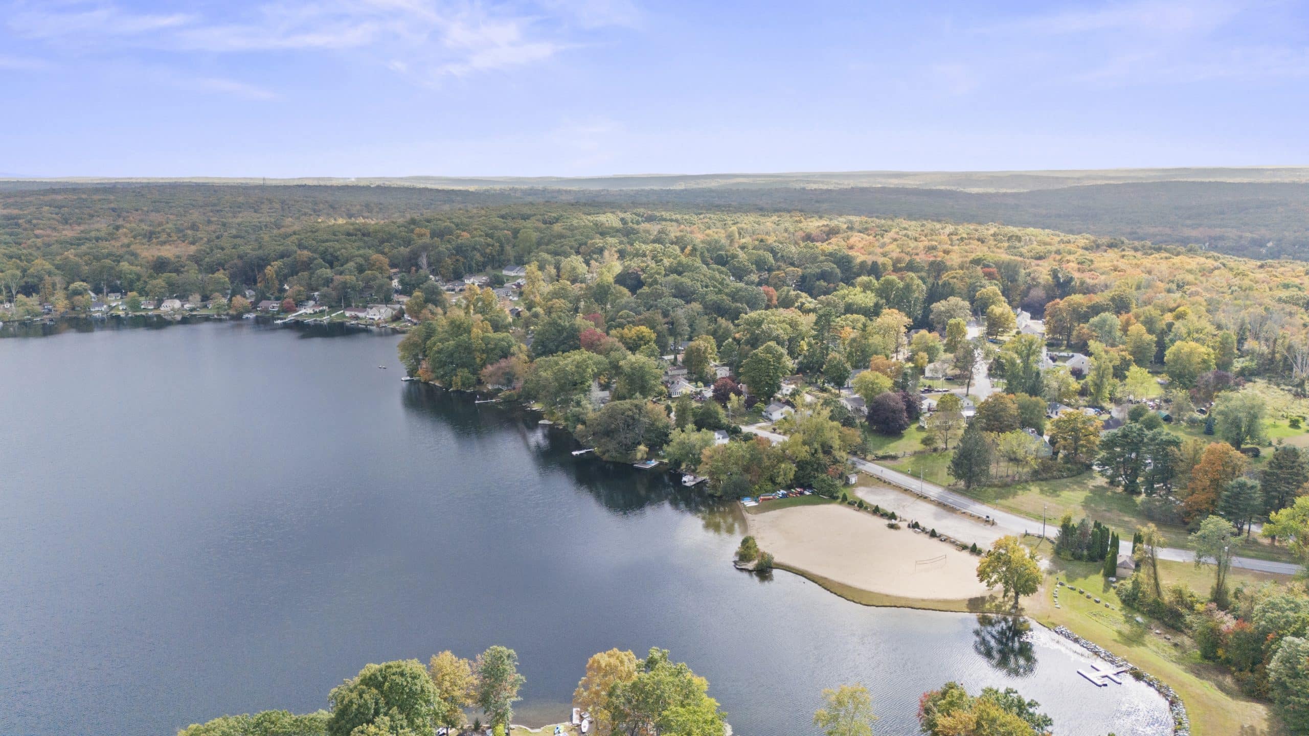 Aerial view of a serene lake surrounded by lush forested hills and scattered houses. A sandy beach area is visible in the foreground, with calm water reflecting the clear blue sky above.