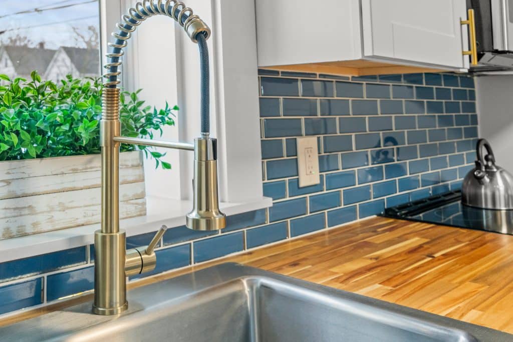 A modern kitchen sink with a stainless steel faucet, wooden countertop, glossy blue subway tile backsplash, and a green plant in a white box on the windowsill.
