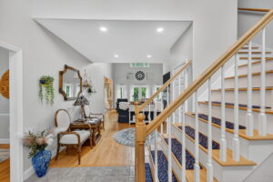 A bright, open foyer with a wooden staircase, a console table with chairs and a mirror, hardwood floors, and a glimpse into a living area with large windows and a decorative wall clock.