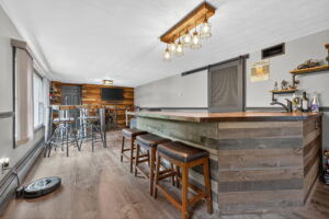 A modern home bar room with a wooden bar counter, three barstools, high-top tables with chairs, a wall-mounted TV, and rustic decor in neutral tones, wood accents, and industrial-style lighting.