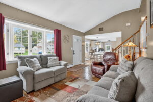 A cozy living room with a gray sofa, armchair, and patterned rug, featuring large front windows with red curtains. The space opens to a dining area and staircase, with natural light brightening the room.