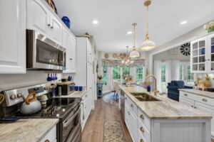 Bright, modern kitchen with white cabinets, stainless steel appliances, marble countertops, gold pendant lights, and a large center island. In the background, a cozy dining area and living space with large windows are visible.