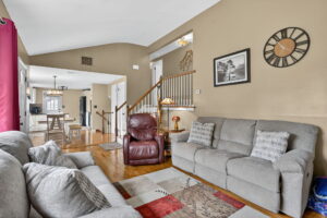 A cozy living room with tan walls, gray sofas, a red recliner, a patterned rug, wall clock, framed photo, and a view into a dining area and kitchen. Stairs with a wooden railing lead to the upper floor.