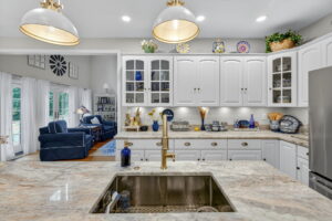 Bright, elegant kitchen with marble countertops, a gold faucet, white cabinets, and blue-and-white decor. Open layout reveals a cozy living room with blue furniture, a large clock, and natural light from French doors.