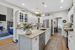 Bright, spacious kitchen with white cabinets, marble countertops, stainless steel appliances, gold pendant lights, and decorative plants. An island with a sink is centered, and adjoining rooms are visible in the background.