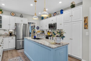 A modern kitchen with white cabinets, stainless steel appliances, a blue island with a marble countertop, gold pendant lights, and various decorative plates and vases displayed above the cabinets.