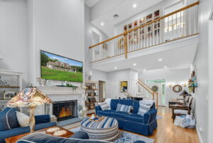 A spacious living room with blue and white furniture, a lit fireplace, a large TV above the mantel, wooden floors, and a loft with a white railing overlooking the space. Stairs lead to the upper level.