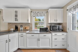 Bright, modern kitchen with white cabinets, granite countertops, and stainless steel appliances. A window with a floral valance lets in natural light; a microwave, toaster oven, and utensils sit on the counters.
