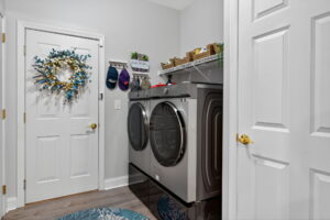 A modern laundry room with a front-loading washer and dryer, shelves with baskets above, a decorative wreath on the door, and hooks holding hats and a Laundry sign on the wall. The floor has a blue-patterned rug.