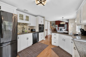 Modern kitchen with white cabinets, black appliances, granite countertops, and a mosaic tile backsplash. Wooden floors, two rugs, and a view into a dining area with red accent wall and white table are visible.