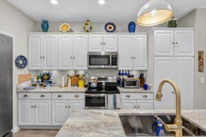 Bright kitchen with white cabinets, marble countertops, stainless steel appliances, and gold fixtures. Blue and yellow decorative items, dishes, and vases accent the space, with under-cabinet lighting and a central island sink.