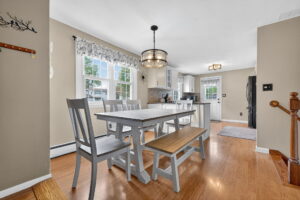 A bright kitchen and dining area with wood floors, a gray wooden dining set with chairs and a bench, white cabinets, stainless steel appliances, and large windows letting in natural light.