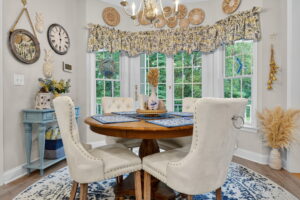 A cozy dining area with a round wooden table, four white upholstered chairs, blue-and-white decor, large windows with floral valances, and natural light illuminating the space. A chandelier hangs above the table.