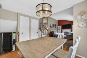 A modern dining area with a light wood table and white chairs, a large round chandelier above, beige walls, white double doors, and a view into a cozy living room with a red accent wall and a fireplace.