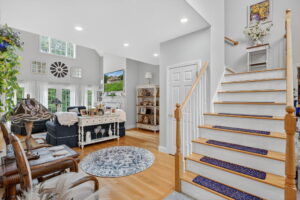 Bright, open living room with wooden floors, light gray walls, and white trim. Stairs with blue runners lead up. Cozy seating, shelves, plants, and decor create a welcoming, airy space with large windows and natural light.