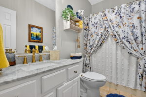 A bright bathroom with white cabinets, a marble countertop, gold fixtures, a toilet, floral shower curtain, blue and yellow towels, decorative shelf with plants, and a framed sunset painting above the towel rack.