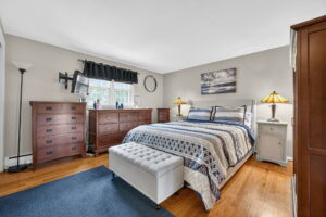 A cozy bedroom with a large bed, patterned bedding, two bedside tables with lamps, a gray tufted bench, wooden dressers, a blue rug, hardwood floors, and a window with a black valance.