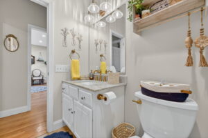 A bright bathroom with a vanity, marble countertop, gold fixtures, a large mirror, wall art, hanging decor, and a toilet. Yellow towels, blue rug, and wood floor add color; a room with a chair is visible through the doorway.