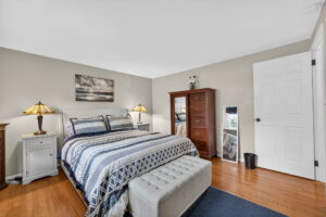 A cozy bedroom with a neatly made bed, patterned blue and white bedding, two nightstands with lamps, a tall wooden dresser, wall art, hardwood floor, and a white door.