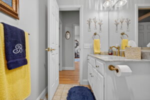A bright bathroom with white cabinets, gold fixtures, yellow towels, a navy blue towel with a B monogram, wall art above the mirror, tissue box, and a view into a hallway with wooden floors.