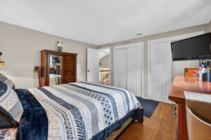 A bedroom with a patterned bed, wooden dresser, TV on a wooden desk, and white double closet doors. The walls are beige, and hardwood flooring is visible. The room appears bright and tidy.