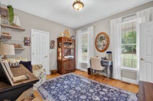 A bright home office with a blue patterned rug, wicker shelving, small desk with a round mirror above, light walls, wooden floor, and large windows with white curtains letting in natural light.