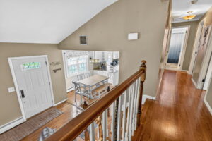 View from a hallway with wooden floors and white railing, overlooking a dining area with a table and chairs near large windows, adjacent to a kitchen; a white front door is visible on the left.