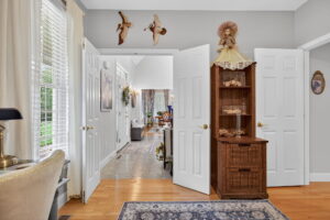 A cozy room with light gray walls, double doors opening to a hallway, a wooden cabinet with a doll on top, decorative birds on the wall, a desk with a chair, and sunlight streaming through large windows with white curtains.