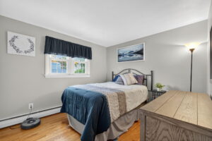 A small bedroom with a neatly made bed featuring blue and beige bedding, a window with a dark curtain, a dresser, a side table with a small plant, wall art, a floor lamp, and a robotic vacuum on the hardwood floor.