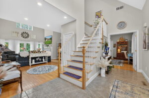 Bright, open living area with wooden staircase, blue and beige decor, a cozy sitting area, a small office with a desk to the right, and large windows letting in natural light. Light walls and hardwood floors create a welcoming space.