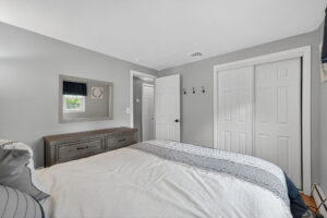 A bedroom with gray walls, a bed with white and blue bedding, a wooden dresser with a mirror above it, a window with a black valance, double closet doors, and an open door leading to a hallway.