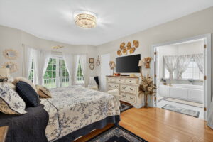 A cozy bedroom with a large bed, floral bedding, light wood floors, a dresser, and a TV. French doors open to greenery outside. An adjoining bathroom with a bathtub and large windows is visible. Decor includes wall art and flowers.