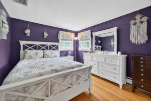A cozy bedroom with purple walls, a white bed with floral bedding, white dresser and mirror, wooden floor, and dreamcatcher wall decor. Two windows with ruffled curtains let in natural light.