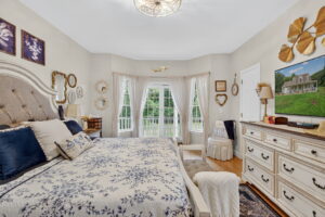A cozy, vintage-style bedroom with a large bed, floral bedding, navy and beige pillows, and a white dresser. The room has framed wall art, gold accents, and a bay window with sheer curtains overlooking greenery.