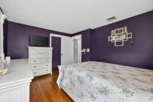 A bedroom with dark purple walls, wooden floor, a white dresser with a TV, a white bed with a floral quilt, and a decorative wall shelf holding framed pictures and small items.