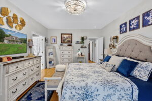 Elegant bedroom with a cream and blue color scheme, featuring a tufted headboard, floral bedding, matching dressers, gold wall decor, framed art, a TV, and a chandelier on the ceiling.