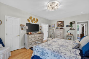 Cozy bedroom with light-colored walls, wood floor, floral bedspread, TV on a dresser, chest of drawers, wall art, decorative items, large mirror, and sunlight streaming through a window on the right.