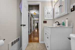 A bathroom with white cabinets and a sink, a mirror, and various toiletries on the counter. The door is open, showing a hallway with wooden floors and ceiling lights.
