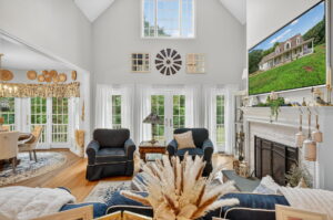 Bright living room with vaulted ceiling, large windows, and a fireplace topped with a TV. Navy armchairs, light décor, and natural accents create an inviting, airy space with views into a dining area.
