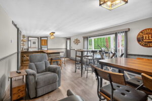 A spacious, bright game room with high tables, mixed seating, a gray recliner, wall decor, a shuffleboard table, and large glass doors leading to a green outdoor area.