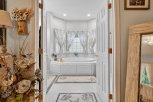 A cozy bathroom with double doors opens to a large white bathtub beneath curtained windows. Floral rugs, a vase with dried flowers, and a wooden-framed mirror accent the inviting, softly lit space.