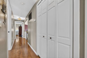 A hallway with wood floors, beige walls, white trim, and double white closet doors. There are doors on both sides and a view into a room with a dresser and lamp at the end of the hall.