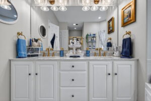 A bathroom with a double sink vanity, gold fixtures, white cabinets, blue towels, and a large mirror. Decorative items and paintings line the walls, and shelves hold various toiletries. The space is bright and neatly organized.
