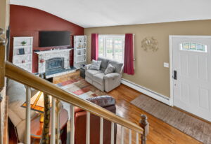 A cozy living room with a red accent wall, white brick fireplace, large TV, bookshelves, gray sofa, leather chair, and area rug. Sunlight streams in through windows with red curtains near the front door.